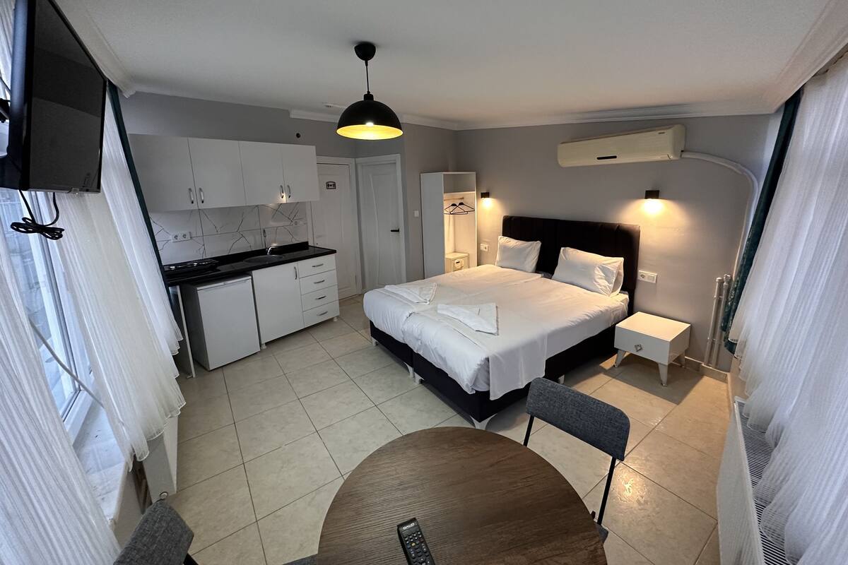A modern hotel room with a bed, bedside tables, a small kitchen area, and a dining table.