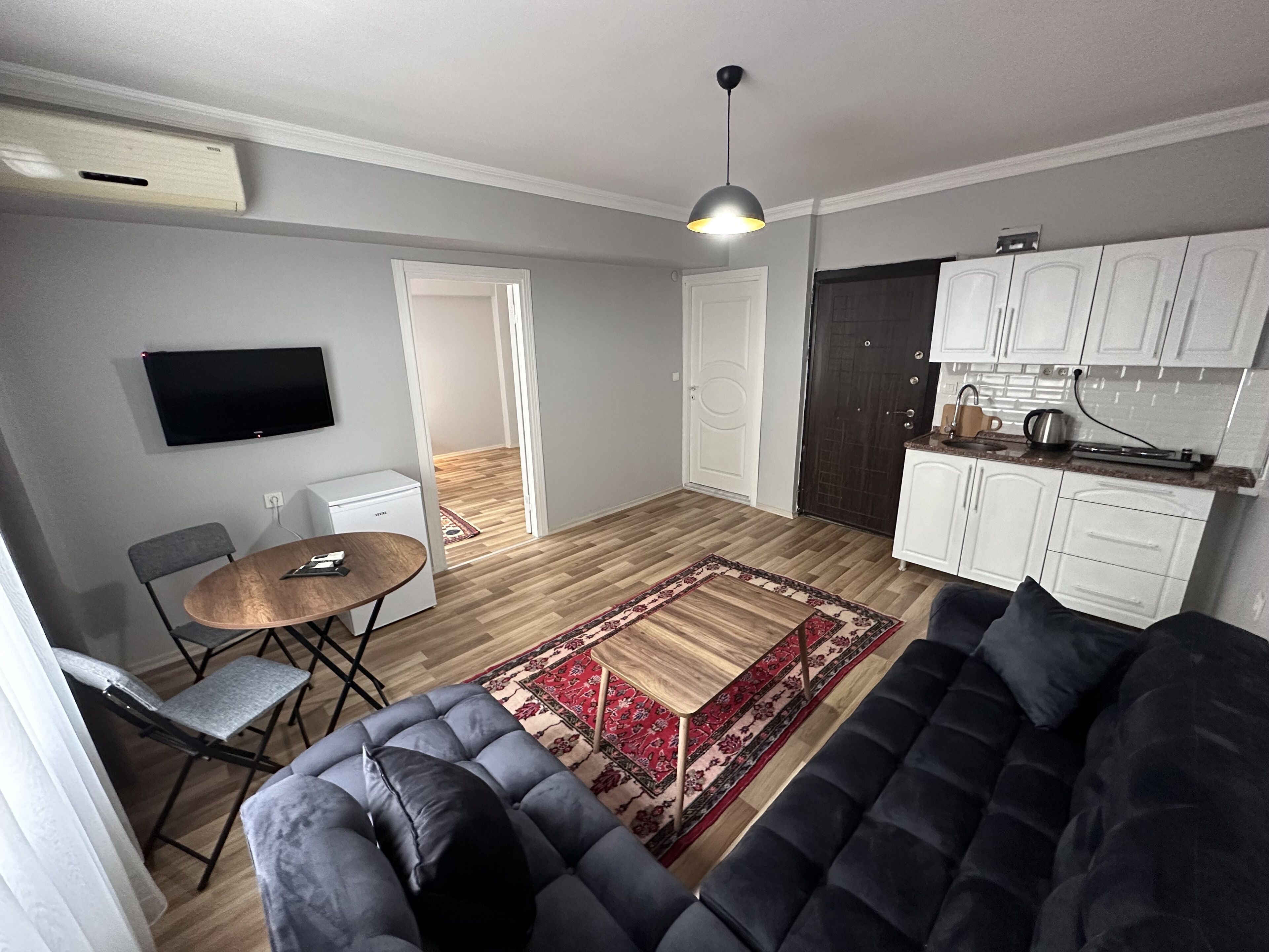 Un espace de vie compact comprenant un canapé, une petite table, un téléviseur, un coin cuisine et une porte donnant sur une autre pièce.