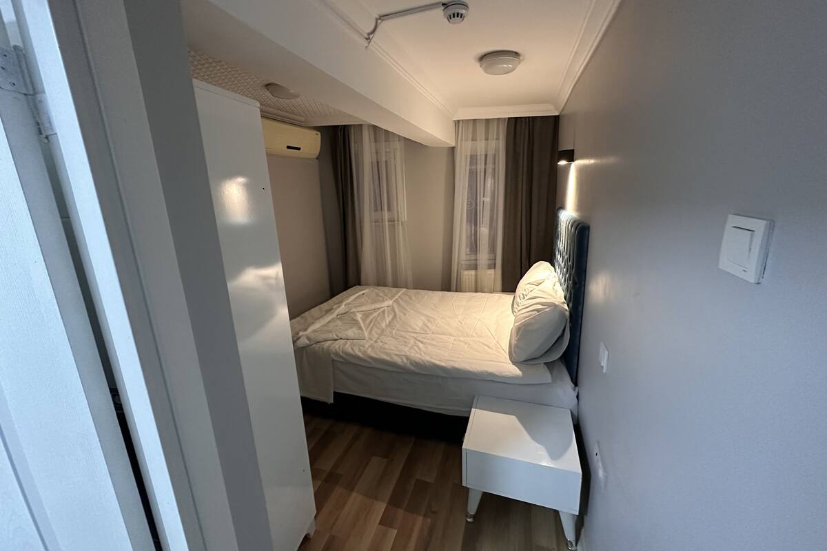 A modern bedroom with a bed, a bedside table, a ceiling-mounted light fixture, and a wall-mounted smoke detector.