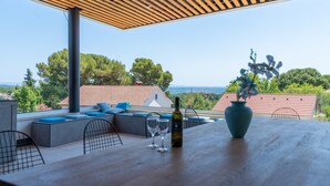 Um terraço com pérgola de madeira, área de estar com almofadas, mesa com garrafa e copos, e vista para o mar e casas.