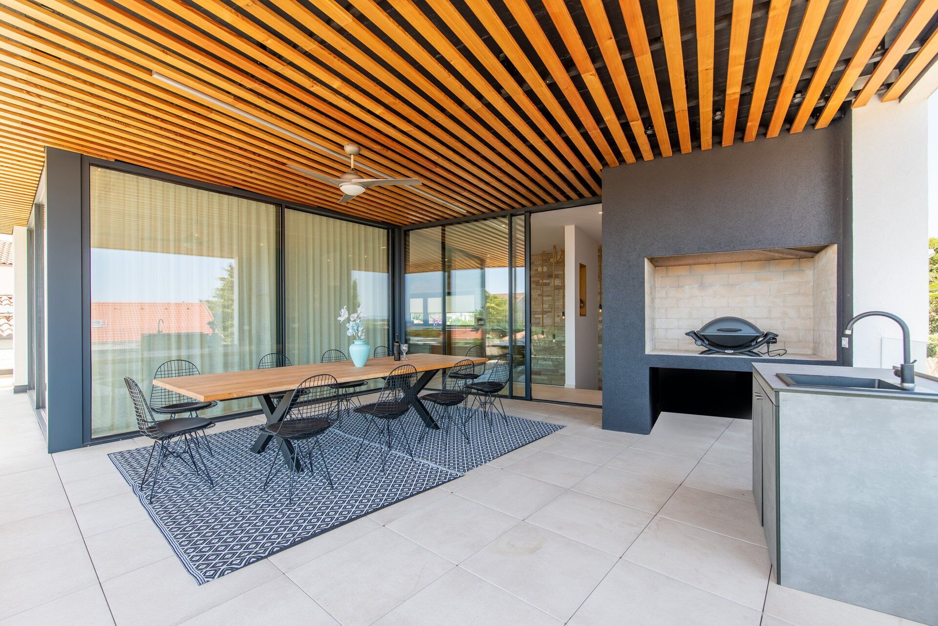 An outdoor patio with a wooden ceiling, a dining table with chairs, a grill, and a kitchen area.