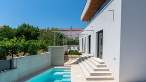 Uma área moderna de piscina ao ar livre, com piscina azul translúcida, degraus que levam a um espaço coberto com assentos, e um edifício branco com telhado em azulejos.