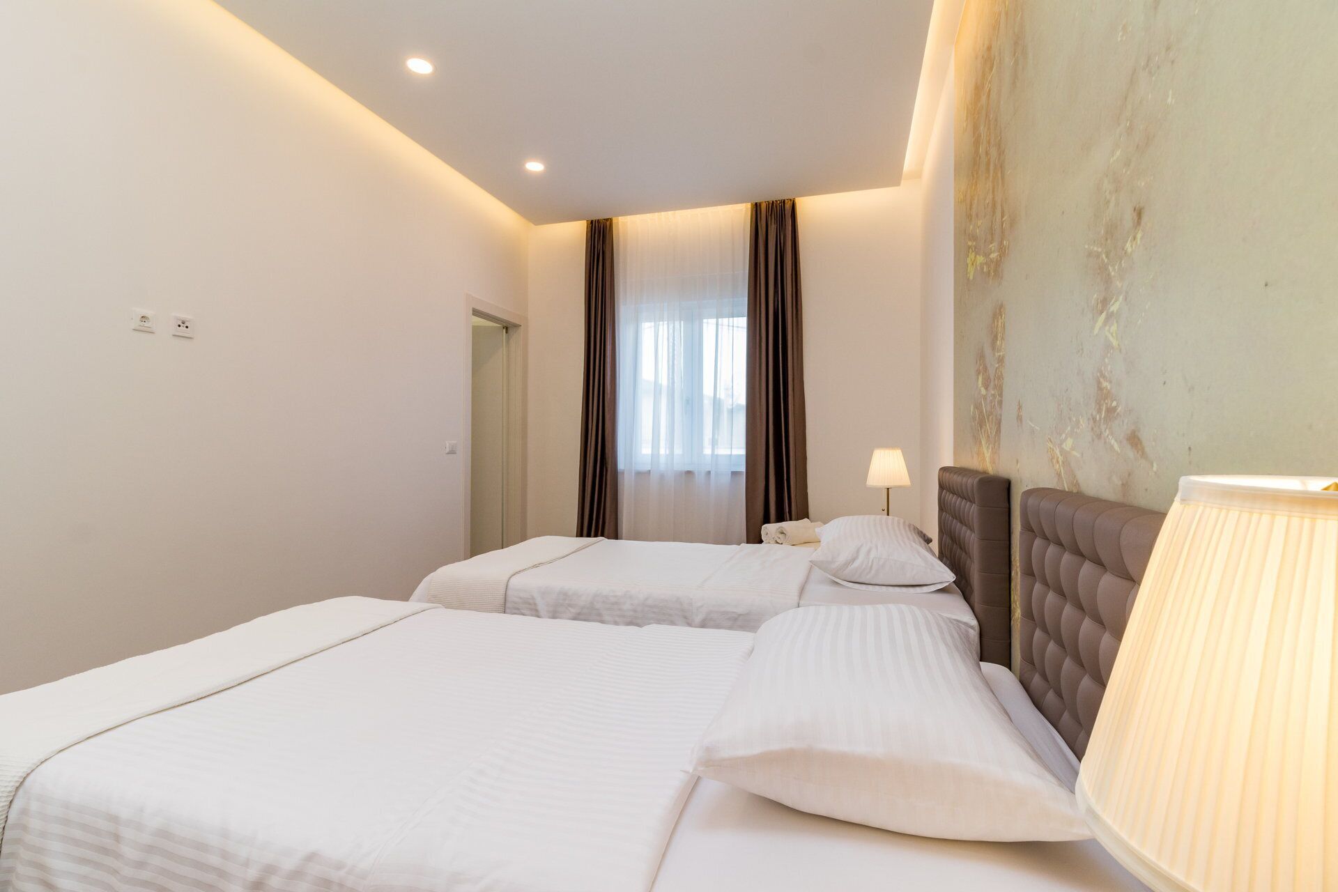 A hotel room with two beds, a bedside lamp, and a view of the outdoors through sheer curtains.