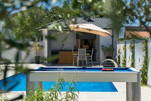 A pool table with a pool in the background, a dining area with chairs, and a covered outdoor seating area.
