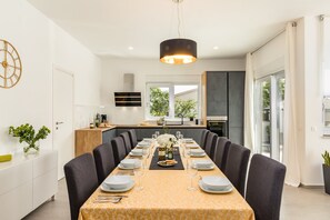 A modern dining room with a long table set for six, a kitchen in the background, and a large window providing natural light.