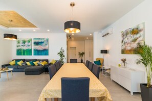 A modern living room with a long dining table, a sofa, and a coffee table. There are two abstract paintings on the wall, a potted plant, and a white cabinet.