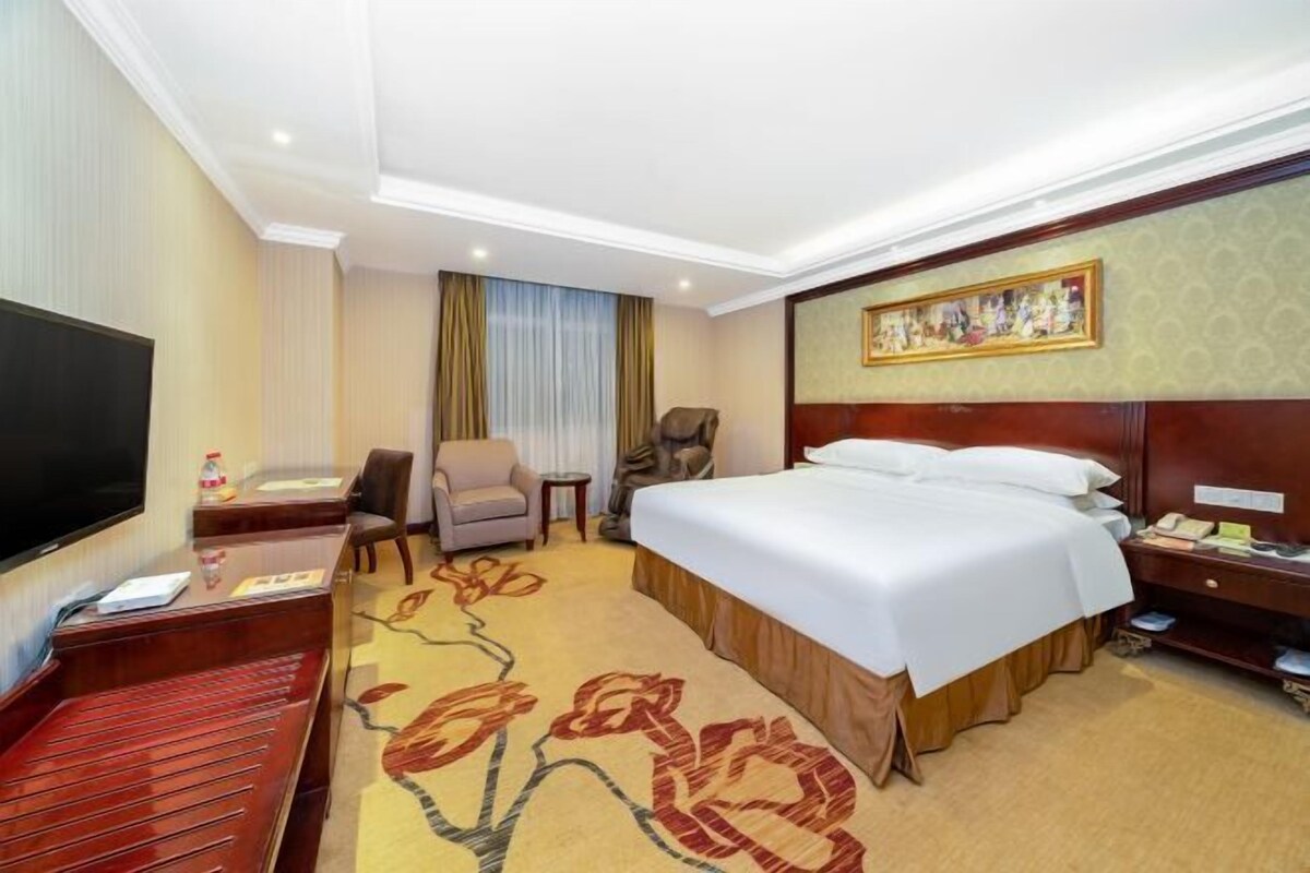 Ein Hotelzimmer mit Bett, Schreibtisch, Stuhl, Fernseher und einem Gemälde an der Wand.