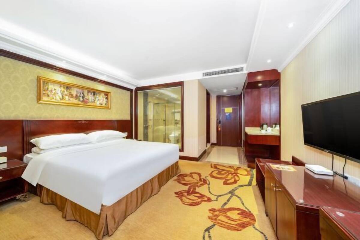 A hotel room with a bed, a television, a desk, and a bathroom visible through a glass door.