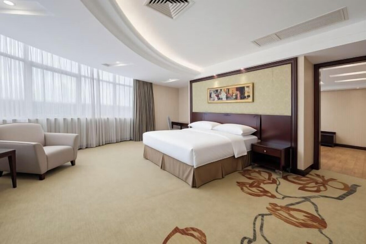 Ein Hotelzimmer mit einem großen Bett, einer Sitzecke, einem Schreibtisch und einem Fenster mit Vorhängen.