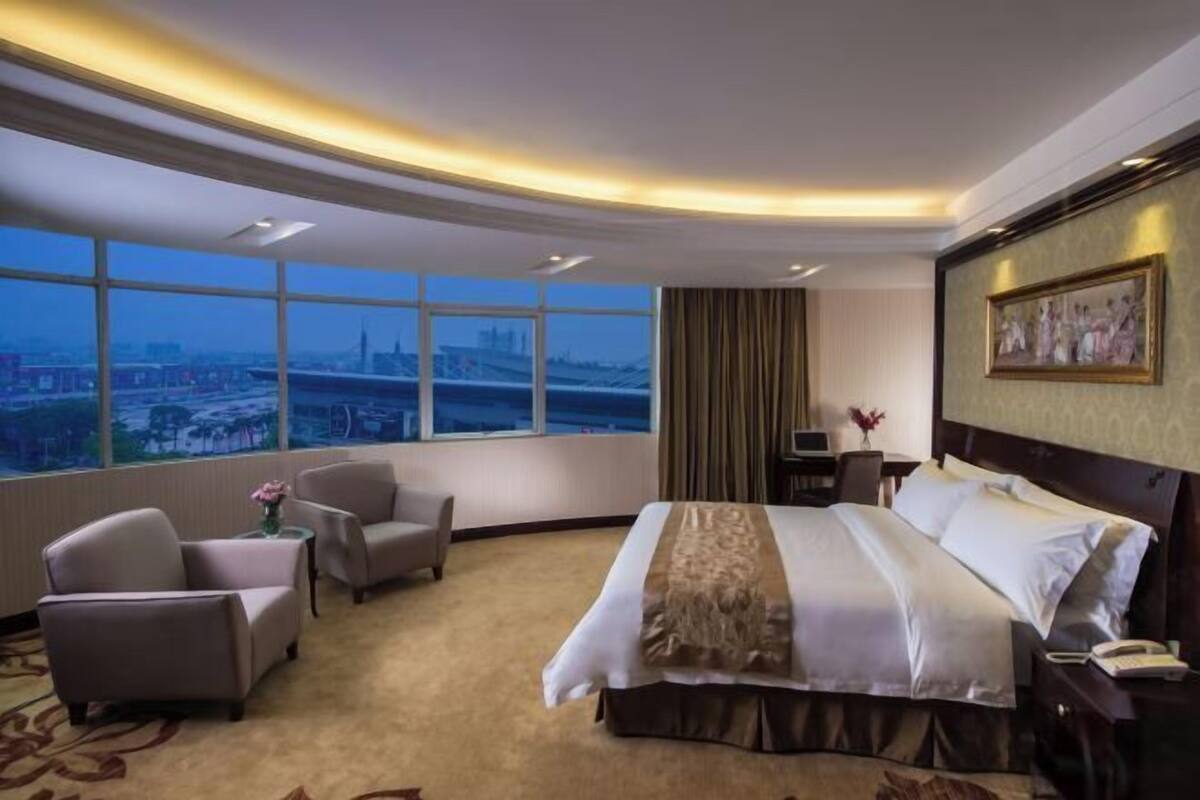 A hotel room with a large bed, two armchairs, a desk, and a view of a cityscape through large windows.