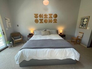 A bedroom with a large bed, two bedside tables with lamps, a chair, a wall art piece, and a macrame wall hanging. - Large Lake House: Expansive View (Paw Paw)