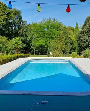 Una piscina rectangular con superficie de agua azul cristalina, rodeada por una plataforma de hormigón y bordeada por un seto bajo.