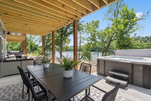 An outdoor patio with a wooden pergola, a dining table with chairs, a hot tub, and a seating area with a sofa and cushions.