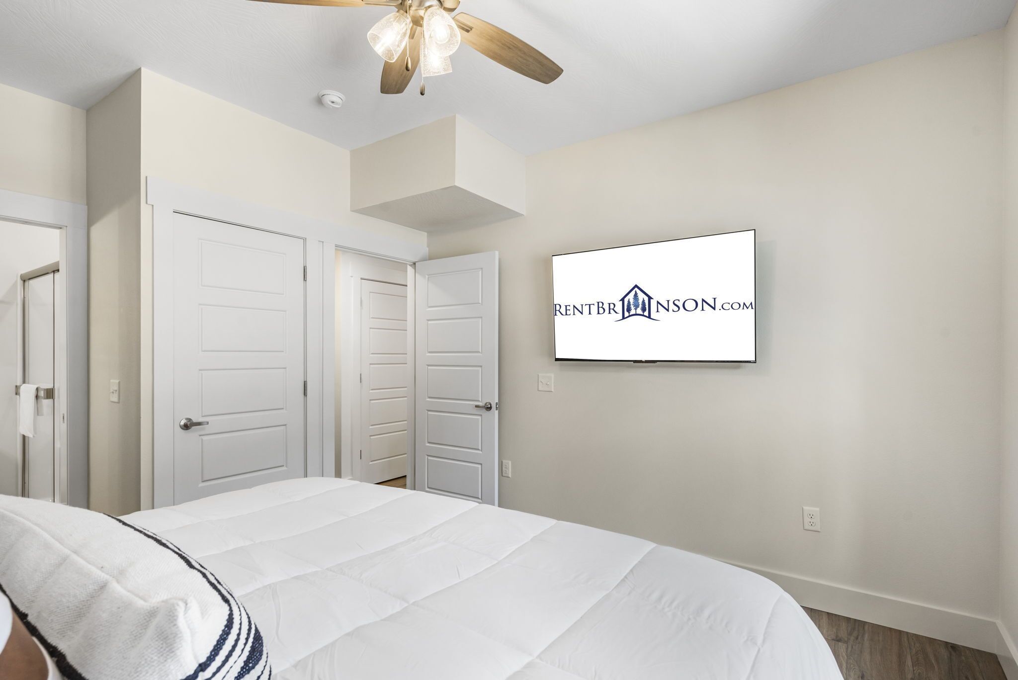 Un dormitorio con cama, dos puertas, un ventilador de techo y un televisor montado que muestra un logotipo.