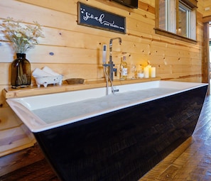 A large, black, freestanding bathtub in a wooden cabin-style bathroom with a wooden wall sign above it.