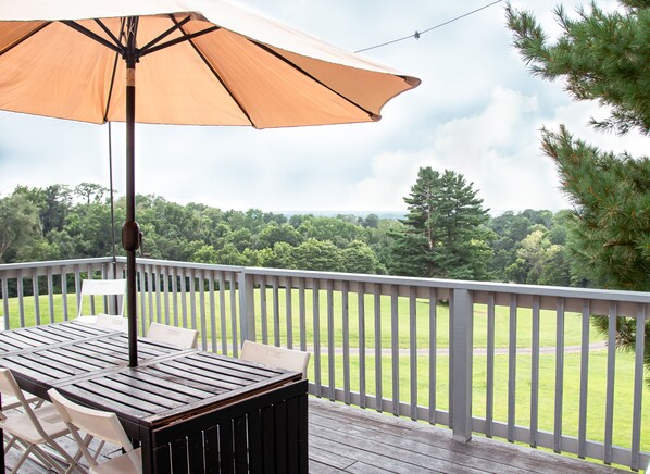 A wooden deck with a table and chairs, an umbrella, and a view of a grassy area with trees.