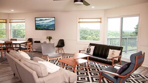Een ruime woonkamer met een bank, stoelen, een salontafel en een eethoek met een tafel en stoelen. Er is een plafondventilator, een schilderij aan de muur en grote ramen met uitzicht op de omgeving.