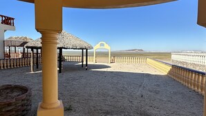 Un espace extérieur abrité, doté d’un sol carrelé, d’un ventilateur de plafond et offrant une vue sur une plage délimitée par une clôture.