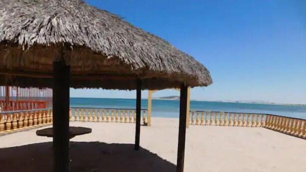 Un abri de plage avec un toit de chaume, des piliers en bois et une plage de sable, avec une rambarde en arrière-plan.