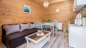 A cozy cabin interior with a dark sofa, a wooden coffee table with fruit, a dining table with chairs, and a kitchen area with a refrigerator and coffee maker.