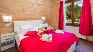 A wooden cabin bedroom with a bed, red curtains, and a window with a view of greenery. - Apartments Plitvice forest - One Bedroom Apartment with Terrace (Plitvicka Jezera)
