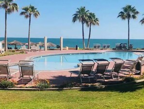 Ein Poolbereich mit Liegestühlen, Palmen und Blick auf das Meer.