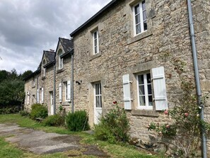Un edificio de piedra con múltiples ventanas y contraventanas, un camino de grava y algo de vegetación.