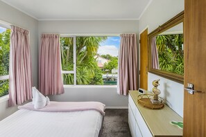 Quarto com cama, mesa de cabeceira com abajur, janela com cortinas e vista para o verde lá fora.