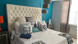 A bedroom with a tufted headboard, a bed with multiple pillows, a bedside table with a lamp, and a window with curtains. - Lovely 3 bedroom condo with pools & gated area. (Fajardo)