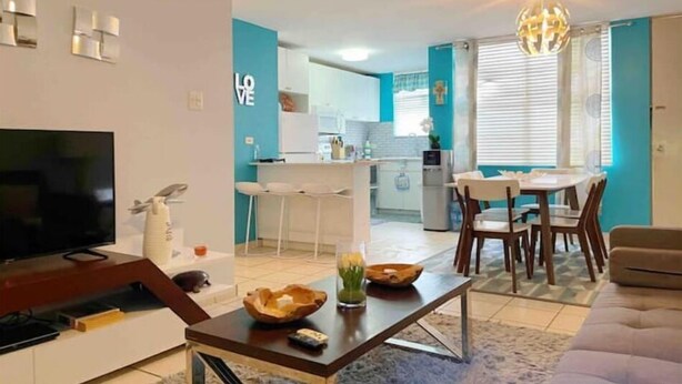 A modern living room with a TV, sofa, and coffee table. A kitchen with a dining area is visible in the background. - Lovely 3 bedroom condo with pools & gated area. (Fajardo)