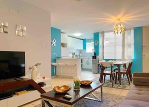 Una sala de estar moderna con televisor, sofá y mesa de centro. Se ve una cocina con comedor al fondo.