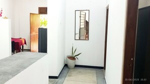 Un pasillo estrecho con suelo de concreto, una puerta de madera a la derecha, una planta en maceta y una mesa roja con una silla al fondo.