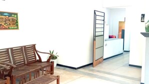 Un pasillo con un banco de madera, una planta en maceta, una pared blanca con un cuadro enmarcado, una puerta con reja metálica y un pilar blanco con un florero.