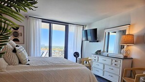 A bedroom with a large bed, a dresser, a TV, and a view of the city. - Oceanfront 11th-Floor End Unit | Quiet & Private | Walk to Barefoot Landing (Myrtle Beach)