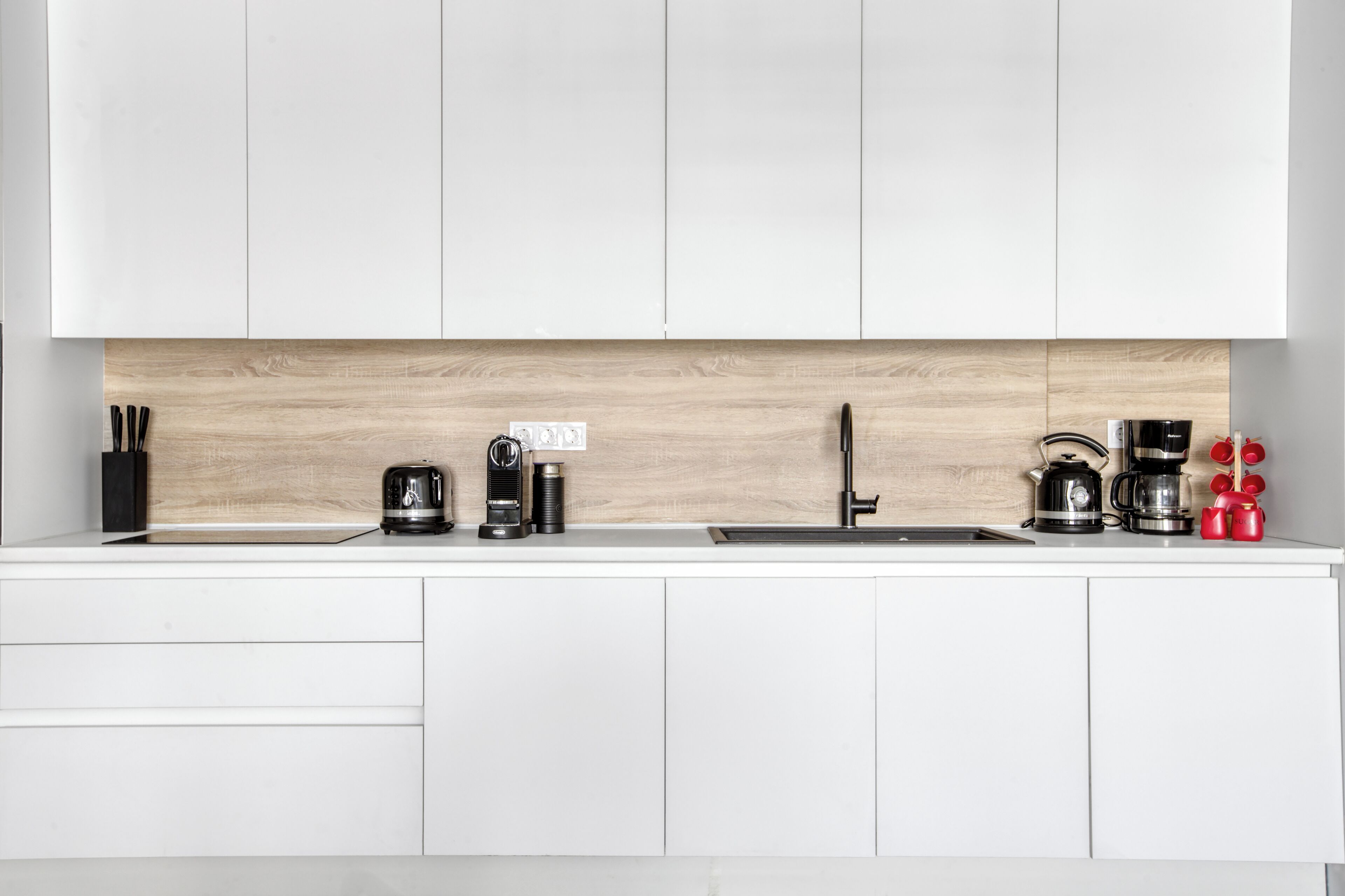 Una cocina moderna con armarios blancos, salpicadero de madera y electrodomésticos de acero inoxidable, incluyendo una cafetera y un hervidor.