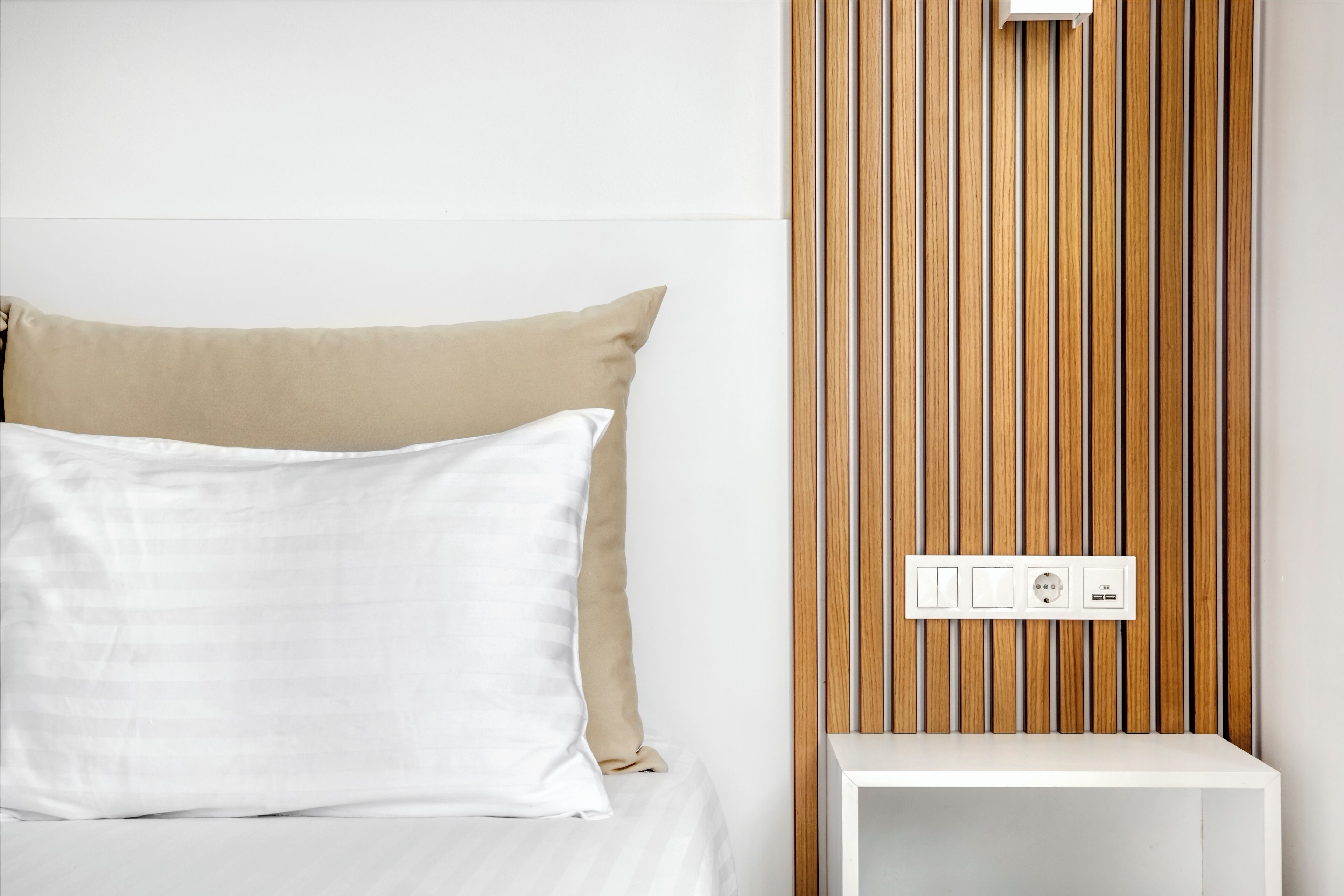 A bed with white and beige pillows, a wooden panel wall, and a white bedside table with a power outlet.
