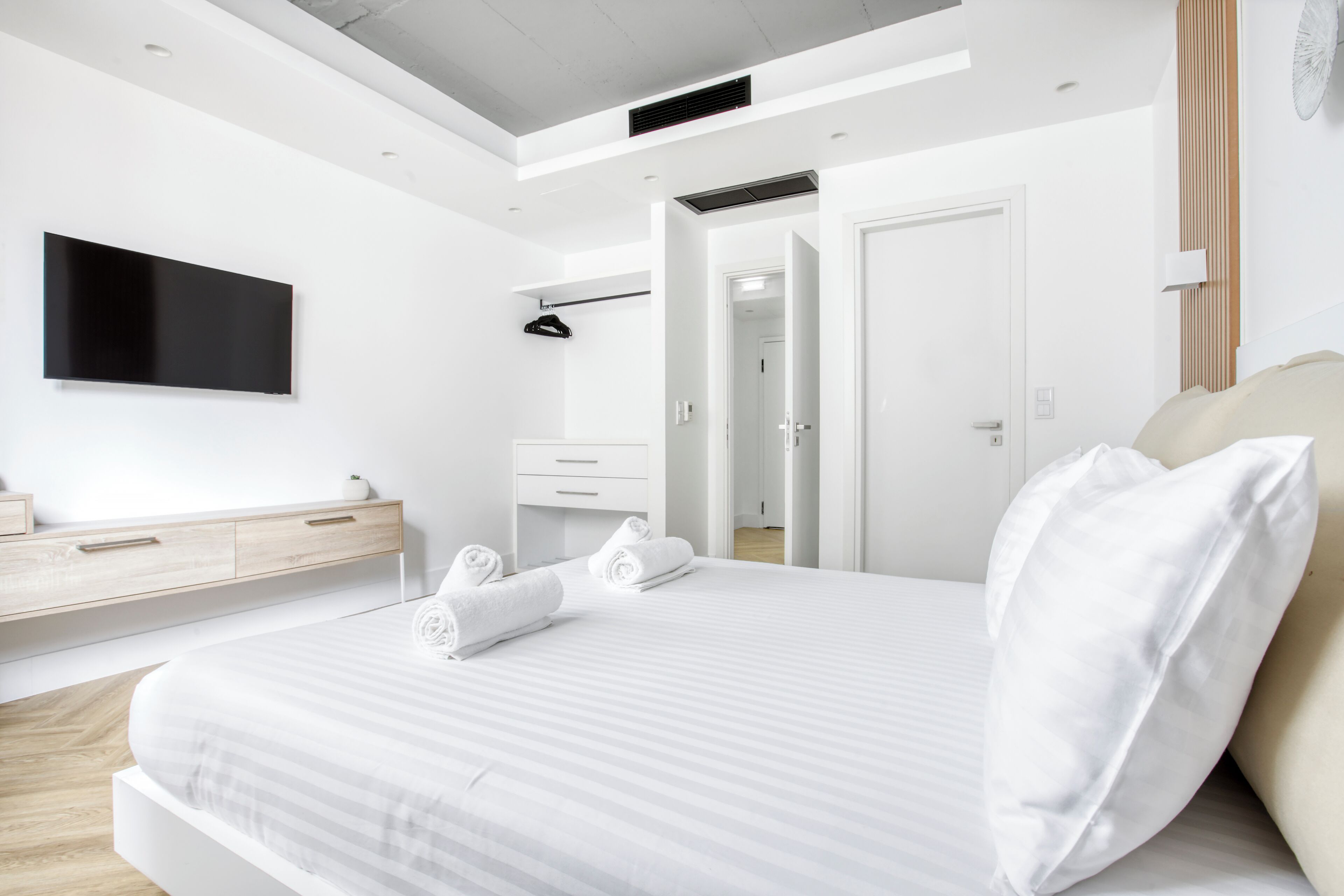 A modern bedroom with a large bed, a flat-screen TV mounted on the wall, a walk-in closet, and a door leading to another room.