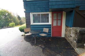 Un petit bâtiment avec une porte rouge, une fenêtre et une table avec deux chaises à l’extérieur.