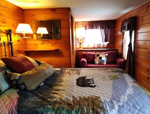 Une pièce chaleureuse avec un décor intérieur en bois, un lit recouvert d’une courtepointe, un canapé rouge et deux lampes.