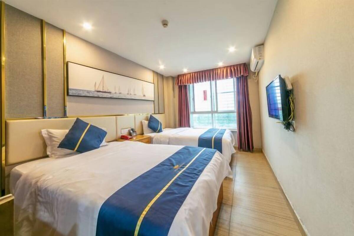 A hotel room with two beds, a large window with curtains, a mounted TV, and a wall-mounted air conditioner.