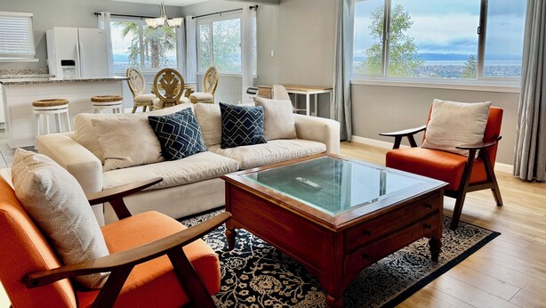 A modern living room with a sofa, coffee table, and chairs. There is a kitchen area with a refrigerator and a dining table. Large windows provide a view of the outdoors. - Spectacular View Attached 2-Car Garage (San Leandro)