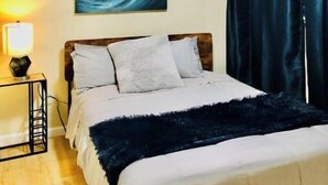 A hotel room with a bed, a nightstand with a lamp, a painting of a wave, and dark curtains. - Great View Guest Unit/Near Eden Hospital/Kaiser SL (San Leandro)