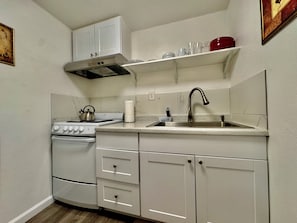 A compact kitchen with white cabinets, a stove, a sink, and a red pot on a shelf.