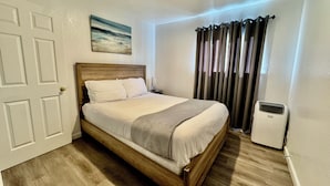 Uma cama bem arrumada com cabeceira de madeira, mesa de cabeceira com abajur, um quadro na parede e uma janela com cortinas.