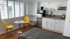 Cocina compacta con refrigerador, microondas y hervidor. Hay un pequeño comedor con una mesa y dos sillas. La habitación tiene un ventanal grande con persianas.