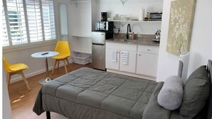 Una habitación compacta con cama, una pequeña mesa con sillas, kitchenette y ventana con persianas.