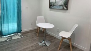 Een kamer met een blauwgroen gordijn, twee witte stoelen, een kleine ronde tafel en een ingelijst kunstwerk aan de muur.