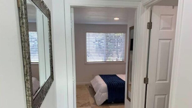 Un couloir étroit mène à une chambre à coucher qui contient un lit, une fenêtre avec des stores et une porte.