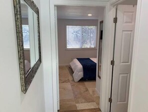 Corredor estreito que dá para um quarto com cama, janela com persianas e uma porta.