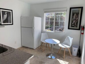 Une cuisine compacte avec un réfrigérateur blanc, deux chaises blanches disposées autour d’une petite table ronde et une fenêtre donnant sur un paysage verdoyant.